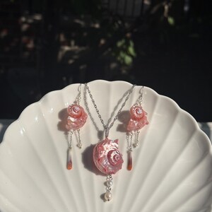Puede incluir: Un conjunto de joyer&iacute;a que consta de un collar y dos pendientes a juego. Cada pieza presenta un componente de concha marina rosa iridiscente y pulida. El collar incluye una cadena de plata, mientras que los pendientes tienen ganchos de plata y peque&ntilde;as perlas blancas.