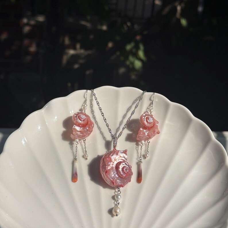 Puede incluir: Un conjunto de joyer&iacute;a que consta de un collar y dos pendientes a juego. Cada pieza presenta un componente de concha marina rosa iridiscente y pulida. El collar incluye una cadena de plata, mientras que los pendientes tienen ganchos de plata y peque&ntilde;as perlas blancas.