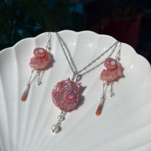Puede incluir: Un conjunto de joyer&iacute;a compuesto por un collar y dos pendientes a juego. Cada pieza presenta un dije de concha espiral rosa iridiscente, acentuado con peque&ntilde;as perlas blancas, cadenas plateadas y colgantes de piedra delgados y c&oacute;nicos.