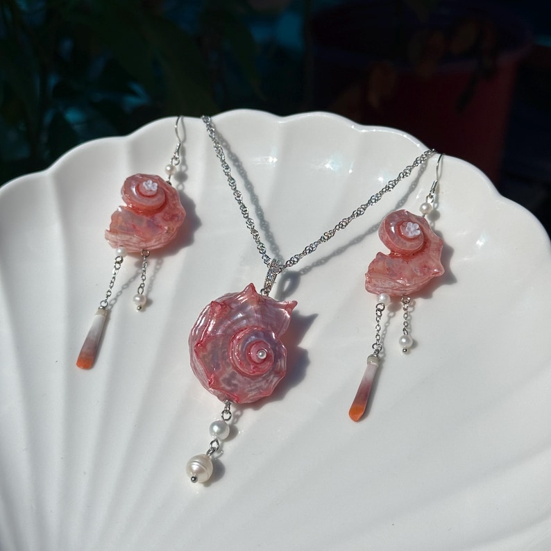 Puede incluir: Un conjunto de joyer&iacute;a compuesto por un collar y dos pendientes a juego. Cada pieza presenta un dije de concha espiral rosa iridiscente, acentuado con peque&ntilde;as perlas blancas, cadenas plateadas y colgantes de piedra delgados y c&oacute;nicos.