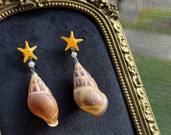 以下が含まれることがあります： 鮮やかな黄色のヒトデのチャームが小さな白いビーズに取り付けられ、茶色とクリーム色の縞模様の自然な螺旋状の貝殻につながっているドロップイヤリングのペア。