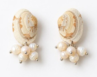 以下が含まれることがあります： ベージュと白の模様が入った天然の貝殻をトップに使用したイヤリングのペア。それぞれの貝殻のベースには、小さな光沢のある白い淡水パールが房状にぶら下がっており、シルバー調の金属パーツで固定されています。