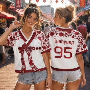 May include: A white short-sleeved jersey with a V-neck, featuring red floral patterns and a red bow graphic on the front. The back displays the name Taehyung and the number 95 in red lettering. The garment includes red trim with geometric designs.