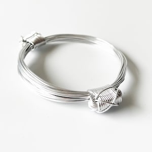 以下が含まれることがあります： 結び目のデザインが施されたシルバーワイヤーのブレスレット。複数のワイヤーを編み合わせて、ユニークでスタイリッシュな外観に仕上げています。