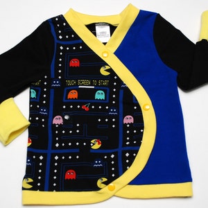 May include: A long-sleeved baby wrap shirt featuring a Pac-Man video game design. The shirt has a black background with a Pac-Man maze pattern, blue and yellow accents, and the text "TOUCH SCREEN TO START".