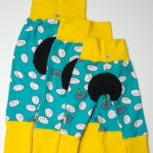 Puede incluir: Tres conjuntos de pijamas infantiles. Los pijamas tienen un fondo turquesa con estampados de nubes blancas y nubes de tormenta grises. Cada conjunto tiene cuello y puños amarillos, y un parche negro en las rodillas.