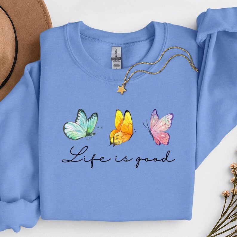 May include: A light blue crewneck sweatshirt featuring a graphic of three watercolor-style butterflies in shades of teal, yellow, and pink. Below the butterflies, the text Life is good is printed in black script. A gold star pendant necklace rests on the collar.