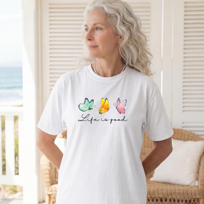 May include: A white short-sleeved t-shirt featuring a graphic of three butterflies in teal, yellow, and pink. Below the butterflies, the text "Life is good" is printed in a black cursive script font. The shirt has a crew neckline and a relaxed fit.