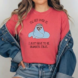 Puede incluir: Camiseta de color coral con un mapache envuelto en una manta azul. El texto encima del mapache dice "I'll get over it." Debajo del mapache, el texto dice "I just have to be dramatic first."