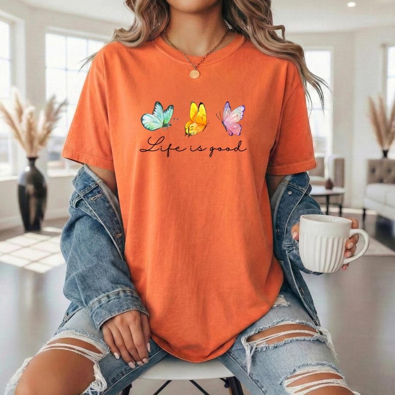 May include: An orange short-sleeved t-shirt featuring three butterflies in teal, yellow, and pink, with the black cursive text Life is good printed below them. The shirt is worn under a blue denim jacket with distressed light blue jeans.
