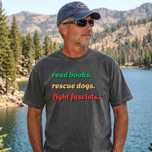 Puede incluir: Una camiseta gris oscuro con el texto "read books. rescue dogs. fight fascists." en verde, amarillo y rojo. La persona de la imagen lleva una gorra azul y gafas de sol. El fondo muestra un lago y montañas.