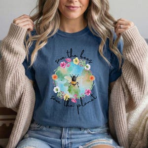 May include: A blue t-shirt featuring a graphic of a bee inside a floral wreath, with the text "Save the bees, save the planet." The design includes a globe and colorful flowers. The shirt is made of soft material.