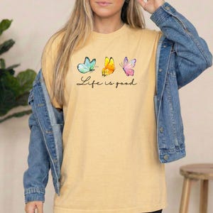 May include: A mustard yellow short-sleeve t-shirt featuring a graphic of three butterflies in teal, yellow, and pink, with the black cursive text Life is good printed underneath. The shirt is worn under a blue denim jacket with metal buttons.