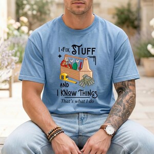 May include: Light blue t-shirt with the text "I Fix Stuff and I Know Things". The graphic includes a wooden toolbox with tools, a measuring tape, and work gloves. The shirt is a crew neck.