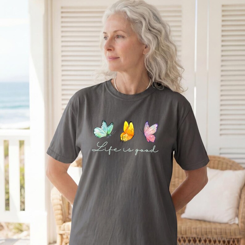 May include: A charcoal gray short-sleeve t-shirt featuring three watercolor-style butterflies in shades of teal, yellow, and pink, with the white cursive text "Life is good" printed directly underneath the butterfly graphic on the front.