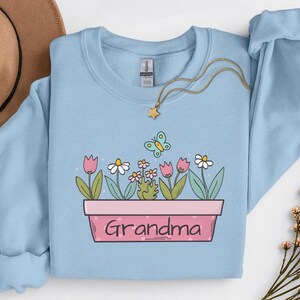 May include: Pale blue sweatshirt with a floral design in a pink planter, the word "Grandma" is written on it. The graphic includes pink tulips, white daisies, and a butterfly. A gold necklace with a star pendant is draped over the sweatshirt.