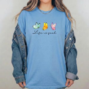 May include: A light blue crew neck t-shirt featuring three watercolor-style butterflies in shades of teal, yellow, and pink, positioned above the black cursive text "Life is good," layered under a blue denim jacket with long sleeves.