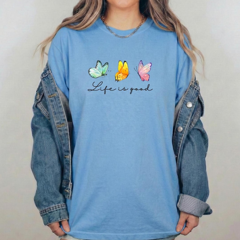 May include: A light blue crew neck t-shirt featuring three watercolor-style butterflies in shades of teal, yellow, and pink, positioned above the black cursive text "Life is good," layered under a blue denim jacket with long sleeves.