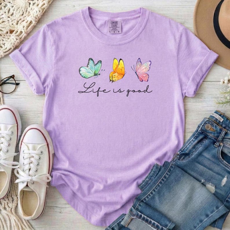 May include: A light purple short-sleeve t-shirt featuring a graphic of three butterflies in blue, yellow, and pink. Below the butterflies, the text Life is good is printed in a black cursive script. The shirt has a crew neckline and a standard fit.