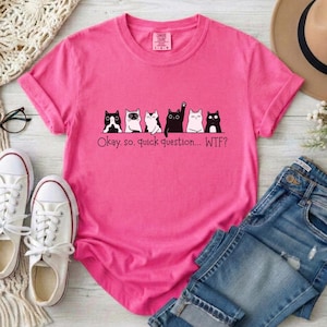 May include: A bright pink short-sleeved t-shirt featuring a graphic of six cats in a row. The text below the cats reads, "Okay. so. quick question... WTF?" The shirt has a crew neckline and a tag at the inner collar area.