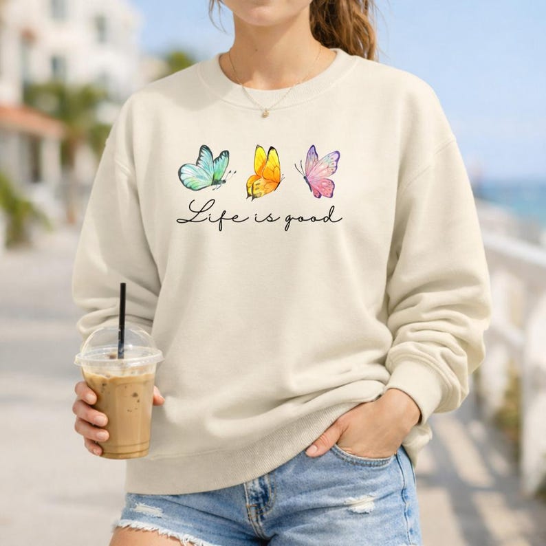May include: A cream-colored crewneck sweatshirt featuring a graphic of three butterflies in blue, yellow, and pink. Below the butterflies, the text "Life is good" is printed in a black cursive script. The sweatshirt has long sleeves and a ribbed collar.