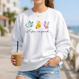 May include: A white long-sleeve crewneck sweatshirt featuring a graphic print of three butterflies in teal, yellow, and pink. Below the butterflies, the text "Life is good" is printed in a black cursive script font across the center of the chest.