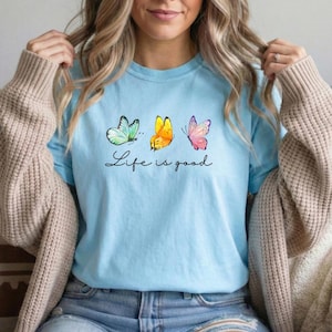 May include: A light blue t-shirt featuring a graphic of three butterflies in teal, yellow, and pink, positioned above the black cursive text Life is good. The shirt has short sleeves and a crew neckline, worn under a beige knit cardigan.