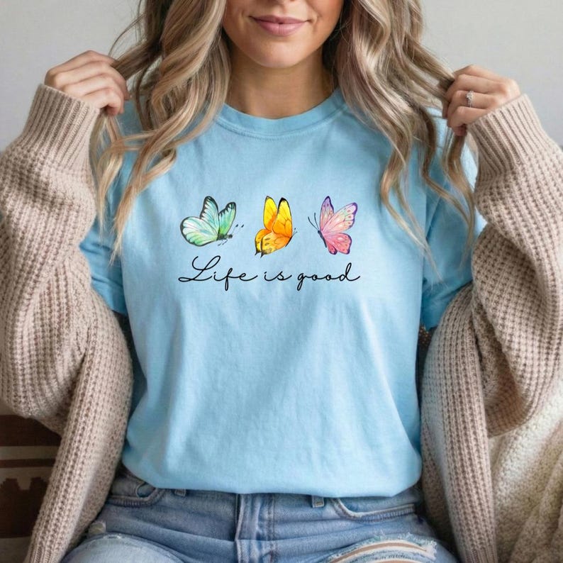 May include: A light blue t-shirt featuring a graphic of three butterflies in teal, yellow, and pink, positioned above the black cursive text Life is good. The shirt has short sleeves and a crew neckline, worn under a beige knit cardigan.