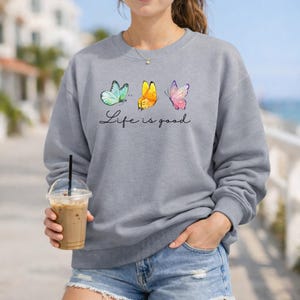 May include: A grey crewneck sweatshirt featuring a graphic of three butterflies in light blue, yellow, and pink. Below the butterflies, the text Life is good is printed in a black cursive script. The sweatshirt has long sleeves and a ribbed neckline.