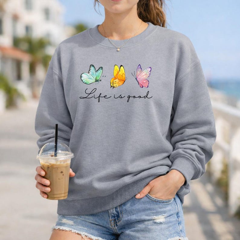 May include: A grey crewneck sweatshirt featuring a graphic of three butterflies in light blue, yellow, and pink. Below the butterflies, the text Life is good is printed in a black cursive script. The sweatshirt has long sleeves and a ribbed neckline.