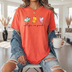 May include: An orange short-sleeved t-shirt featuring three watercolor-style butterflies in blue, yellow, and pink, with the black cursive text Life is good printed underneath. The shirt is worn with a blue denim jacket and distressed blue jeans.