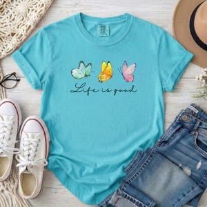 May include: A bright turquoise short-sleeve t-shirt featuring a graphic of three butterflies in light blue, yellow, and pink. Below the butterflies, the text Life is good is printed in a black cursive script. The shirt has a crew neckline.