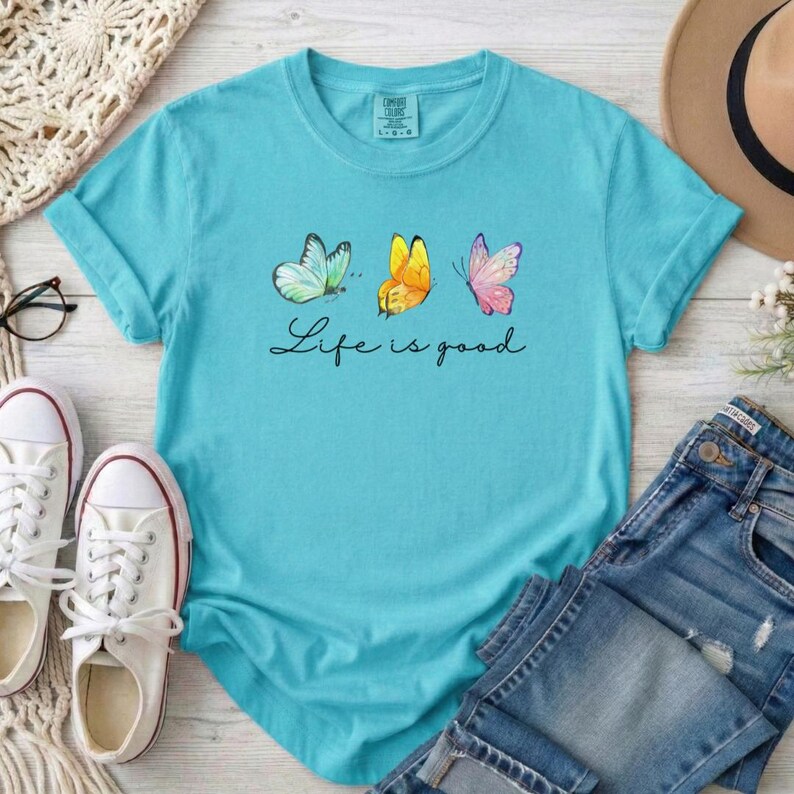 May include: A bright turquoise short-sleeve t-shirt featuring a graphic of three butterflies in light blue, yellow, and pink. Below the butterflies, the text Life is good is printed in a black cursive script. The shirt has a crew neckline.