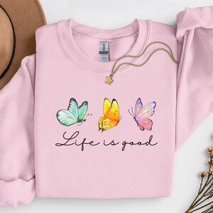 May include: A light pink crewneck sweatshirt featuring a graphic of three butterflies in blue, yellow, and pink, with the black cursive text Life is good printed below. A gold-toned necklace with a small star pendant rests on the neckline.