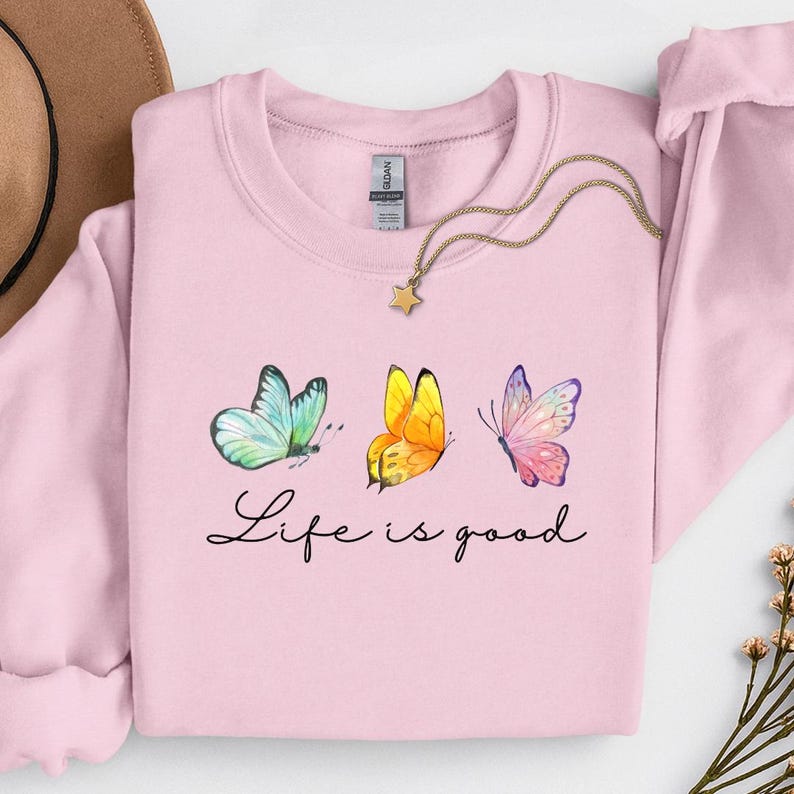 May include: A light pink crewneck sweatshirt featuring a graphic of three butterflies in blue, yellow, and pink, with the black cursive text Life is good printed below. A gold-toned necklace with a small star pendant rests on the neckline.