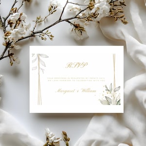 Op de afbeelding: Witte RSVP-kaart met gouden tekst en accenten. De kaart bevat de woorden "RSVP", "Margaret & William" en andere tekst. De kaart is versierd met aquarel bladeren en gouden lijnen. De achtergrond bevat witte stof en bloemtakken.