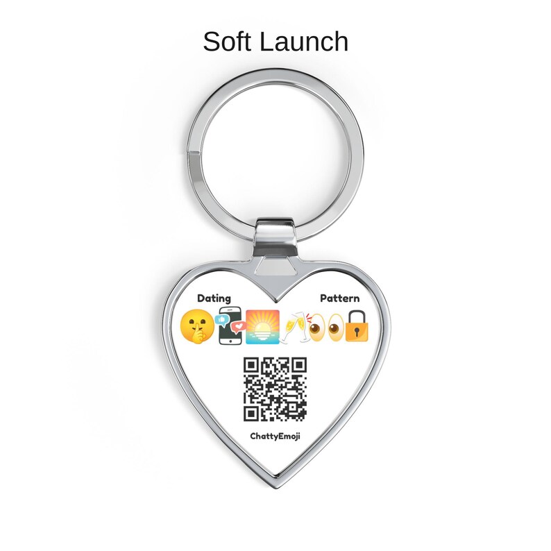 Puede incluir: Un llavero de plata en forma de coraz&oacute;n con el texto Soft Launch en la parte superior. El centro muestra las palabras Dating y Pattern flanqueando una fila de emojis que incluyen una cara pidiendo silencio, smartphone, atardecer.