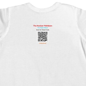 Puede incluir: Una camiseta blanca de manga corta con texto centrado que dice The Nuclear Meltdown, The Public Fallout, Scan for Cheat Code y ChattyEmoji, junto con un c&oacute;digo QR en blanco y negro situado entre las l&iacute;neas de texto.
