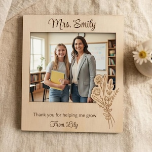 Può includere: Una cornice in legno con una foto di un'insegnante e di una studentessa. La cornice è incisa con le parole "Mrs. Emily", "Thank you for helping me grow" e "From Lily". Un disegno floreale è inciso sul lato destro della cornice.