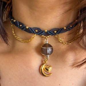Op de afbeelding: Een donkerblauwe macramé choker ketting met gouden accenten. De ketting heeft gouden kralen, een gouden ketting, een grijze stenen hanger en een gouden halve maan met een hart charme. De ketting wordt om de nek gedragen.