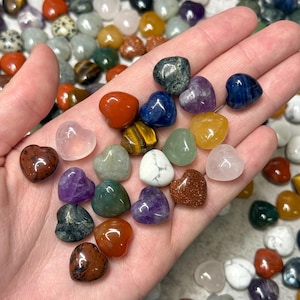 Puede incluir: Una colección de piedras preciosas en forma de corazón de varios colores y texturas. El surtido incluye piedras rojas, azules, verdes, moradas y doradas. Las piedras preciosas son lisas y pulidas, con una variedad de patrones y colores. La imagen muestra una variedad de piedras en forma de corazón.