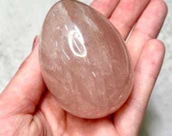 Puede incluir: Un cristal de cuarzo rosa pulido en forma de huevo. La piedra tiene un tono rosa translúcido con sutiles vetas internas. La superficie lisa refleja la luz, resaltando su belleza natural. Un objeto decorativo y único.