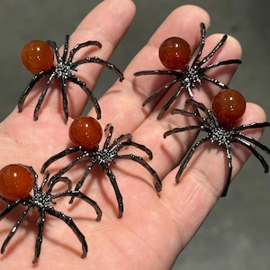 May include: Five decorative spider ornaments. Each spider has a dark gray body and legs, with a round, orange-brown gemstone for the spider's abdomen. The spiders are arranged on a light-colored surface.