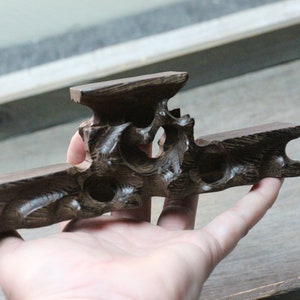 May include: A dark brown, carved wooden display stand with a unique, openwork design. The stand features a horizontal base with a raised platform and a central, decorative element. The wood grain is visible, adding texture to the piece.