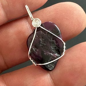 May include: A dark purple, heart-shaped pendant with a silver wire wrap. The wire is intricately coiled at the top for the bail. The stone has a natural, textured surface. The pendant is held in a hand.