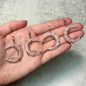 May include: Four rose quartz crescent moon pendants with silver wire wrapping. Each moon has a silver bail for hanging. The pendants are a soft pink color and are displayed on a hand.