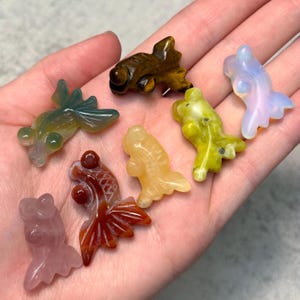 Op de afbeelding: Een verzameling edelstenen goudvisfiguren in verschillende kleuren, waaronder groen, bruin, geel, oranje en roze. Elke figuur is ingewikkeld gesneden met gedetailleerde vinnen en lichamen, die de natuurlijke schoonheid van de stenen laten zien.