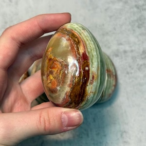 Op de afbeelding: Een gepolijste, eivormige steen met een mix van kleuren, waaronder groen, bruin, rood en geel. De steen heeft een glad, glanzend oppervlak en een natuurlijk uiterlijk. Het ei wordt in een hand gehouden.