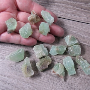 May include: A collection of raw, uncut green calcite crystals. The crystals vary in size and shape, with a translucent, pale green hue and some brown inclusions. The crystals are displayed on a wooden surface.