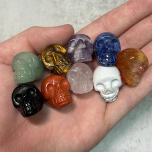 May include: A collection of carved gemstone skulls in various colors, including green, black, red, white, purple, blue, yellow, and orange. Each skull is intricately detailed, showcasing the natural patterns and textures of the stones.